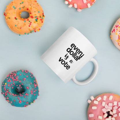 A glossy white ceramic mug with bold, lowercase black sans serif font that reads 'every dollar is a vote', with 'is a' in black handwritten script font, placed on its side against a pastel light blue background. Colorful doughnuts are scattered around the mug, adding a playful touch to the scene.