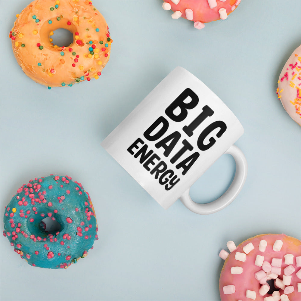 A glossy white ceramic mug with bolded black bubble text that reads 'BIG DATA ENERGY', placed on its side against a pastel light blue background. Colorful doughnuts are scattered around the mug, adding a playful touch to the scene.