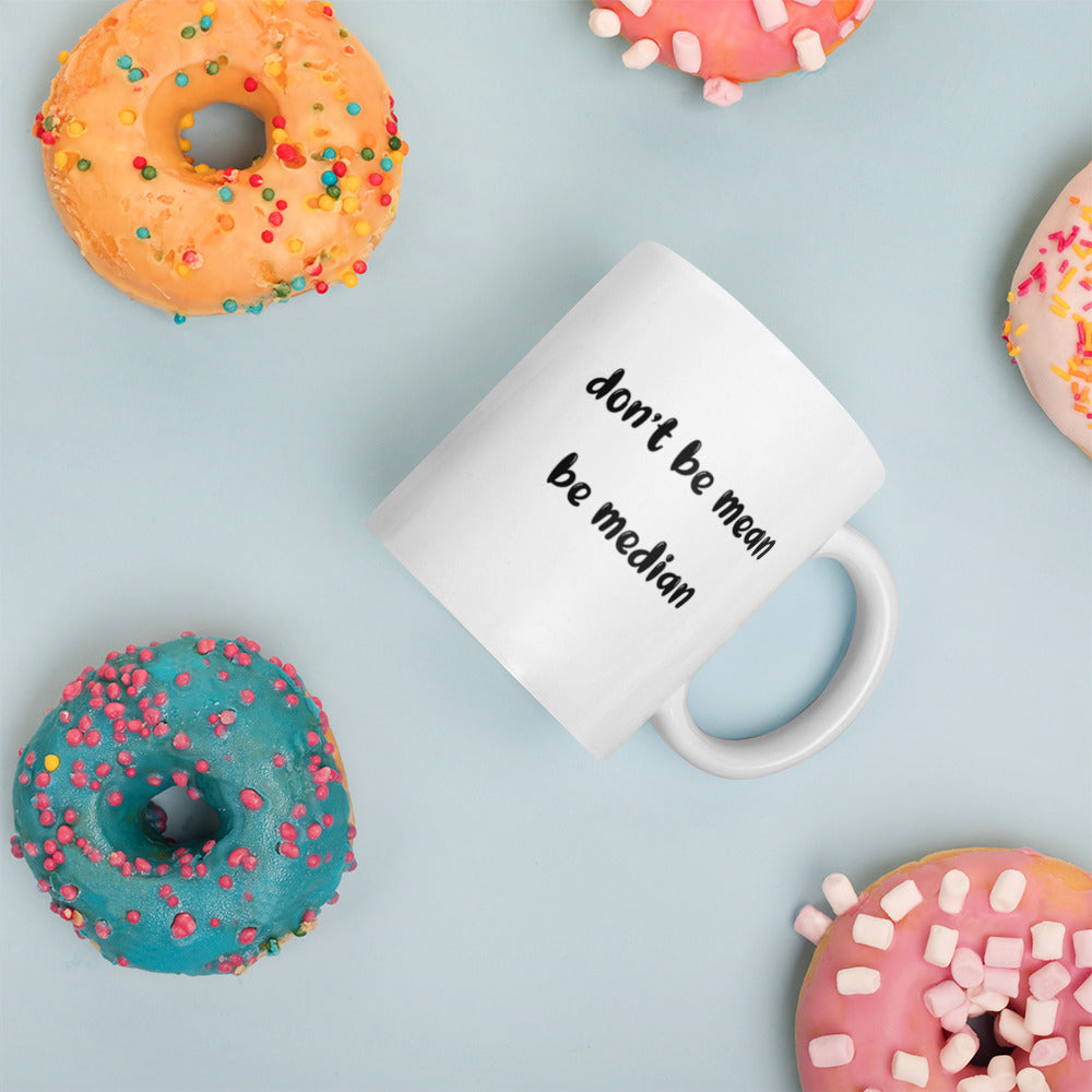 A glossy white ceramic mug with bold, lowercase black bubble font that reads 'don't be mean be median', placed on its side against a pastel light blue background. Colorful doughnuts are scattered around the mug, adding a playful touch to the scene.