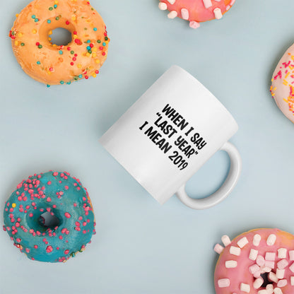 A glossy white ceramic mug with the text 'WHEN I SAY 'LAST YEAR' I MEAN 2019' in black bold uppercase font, placed on its side against a pastel light blue background. Colorful doughnuts are scattered around the mug, adding a playful touch to the scene.