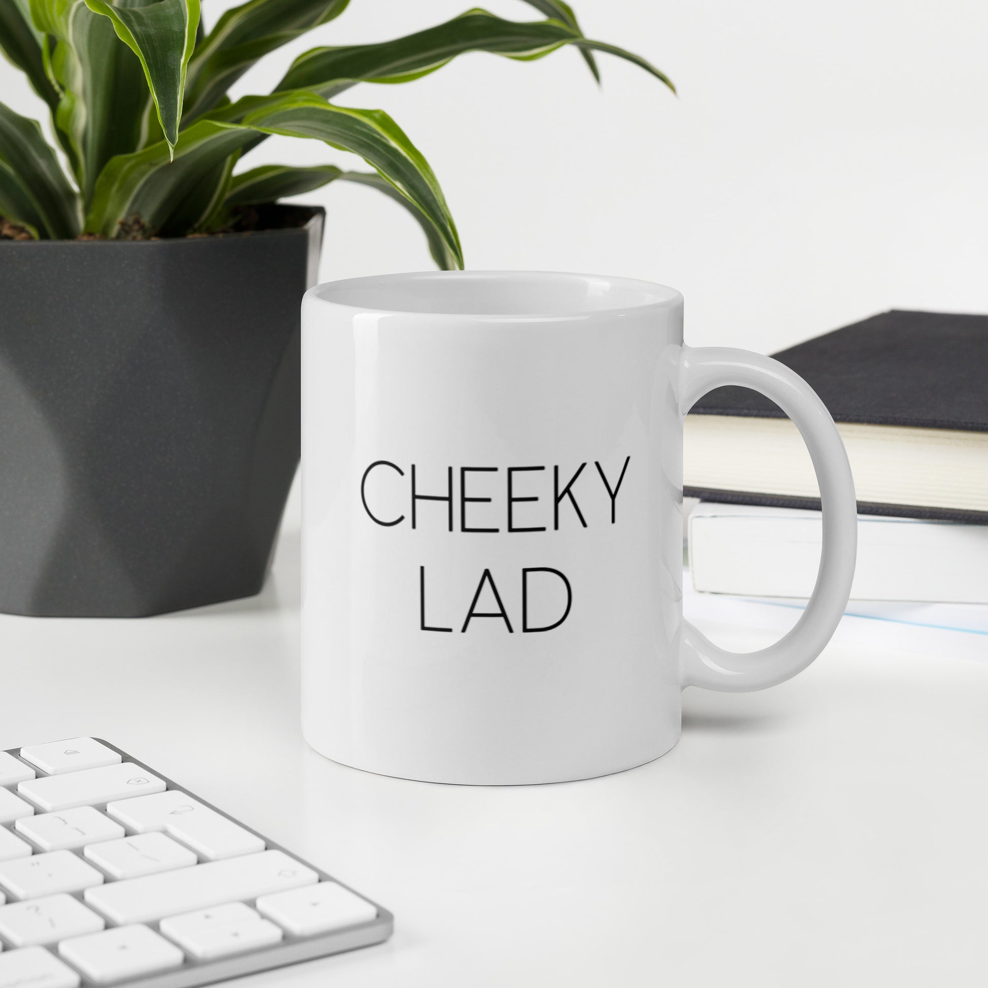 A glossy white ceramic mug with thin black all-caps text that reads 'CHEEKY LAD', sits upright on a white table. In the background, there's a potted plant with downward-curving leaves, resembling a snake plant, in a dark grey pot. Accompanying the plant is a stack of books. A corner of a keyboard is partially visible in the foreground, slightly out of focus, adding a subtle hint of workspace ambiance.