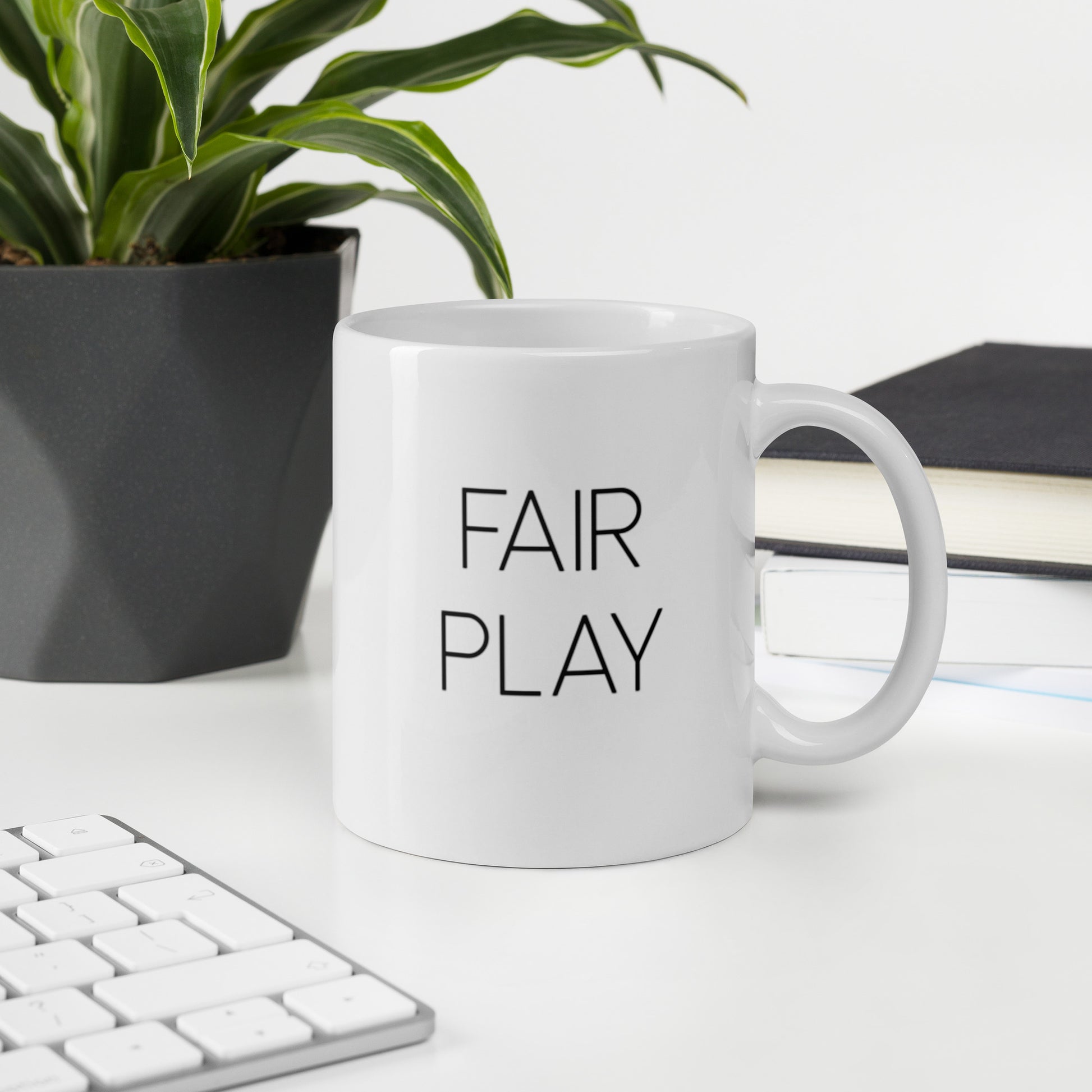 A glossy white ceramic mug with thin, all-caps black sans serif font that reads 'FAIR PLAY', sits upright on a white table. In the background, there's a potted plant with downward-curving leaves, resembling a snake plant, in a dark grey pot. Accompanying the plant is a stack of books. A corner of a keyboard is partially visible in the foreground, slightly out of focus, adding a subtle hint of workspace ambiance.