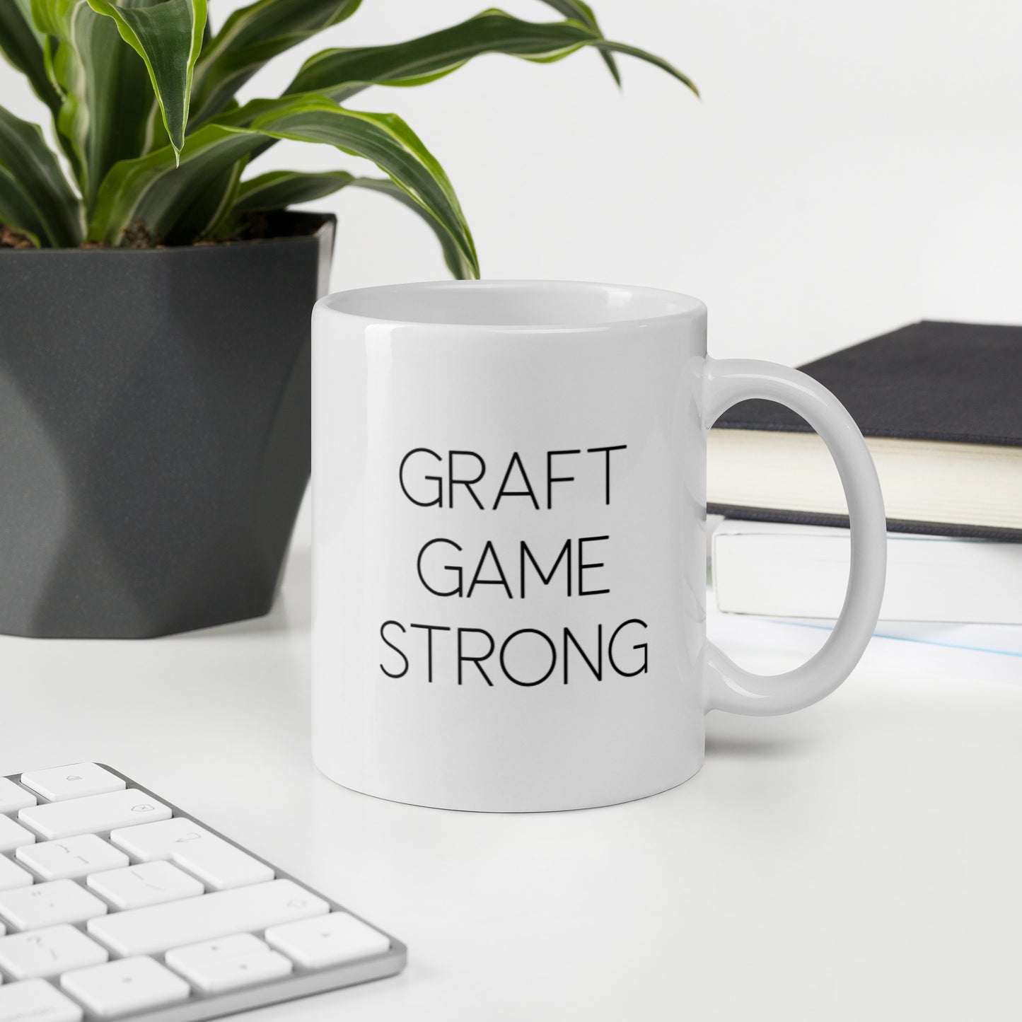 A glossy white ceramic mug with thin, all-caps black sans serif font that reads 'GRAFT GAME STRONG', sits upright on a white table. In the background, there's a potted plant with downward-curving leaves, resembling a snake plant, in a dark grey pot. Accompanying the plant is a stack of books. A corner of a keyboard is partially visible in the foreground, slightly out of focus, adding a subtle hint of workspace ambiance.