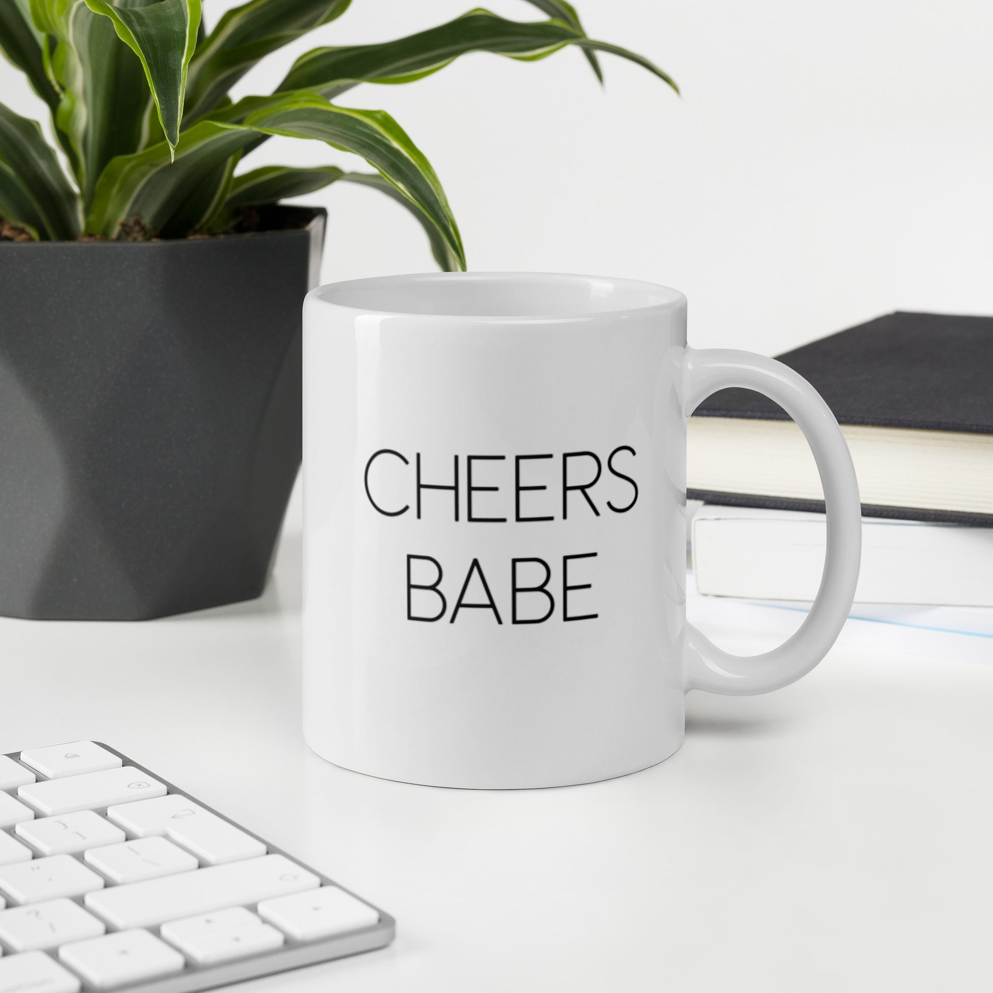 A glossy white ceramic mug with thin black all-caps text that reads 'CHEERS BABE', sits upright on a white table. In the background, there's a potted plant with downward-curving leaves, resembling a snake plant, in a dark grey pot. Accompanying the plant is a stack of books. A corner of a keyboard is partially visible in the foreground, slightly out of focus, adding a subtle hint of workspace ambiance.