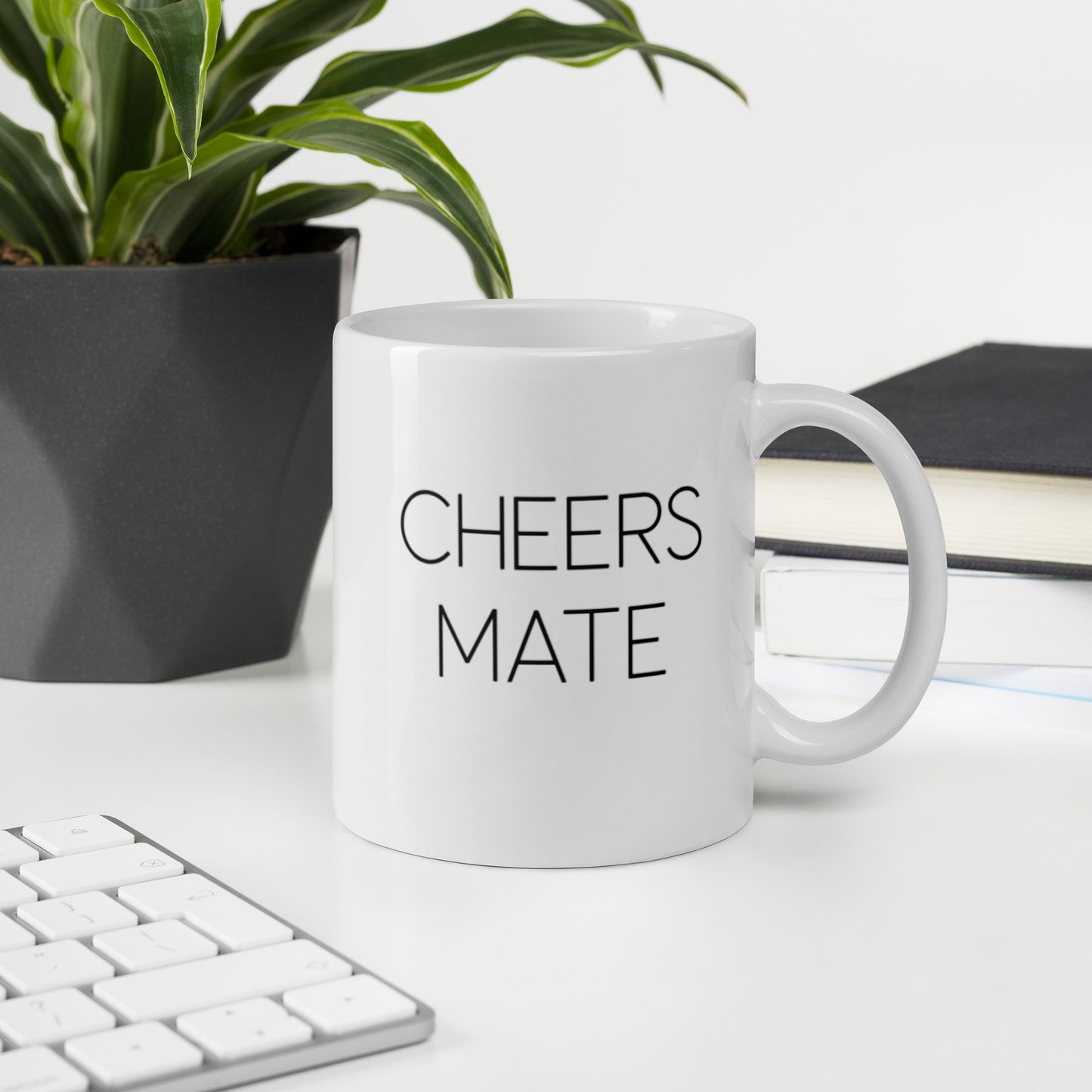 A glossy white ceramic mug with thin black all-caps text that reads 'CHEERS MATE', sits upright on a white table. In the background, there's a potted plant with downward-curving leaves, resembling a snake plant, in a dark grey pot. Accompanying the plant is a stack of books. A corner of a keyboard is partially visible in the foreground, slightly out of focus, adding a subtle hint of workspace ambiance.