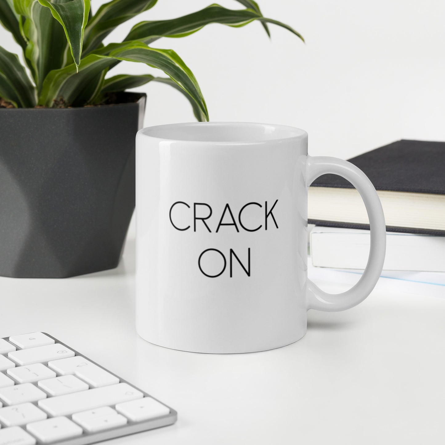 A glossy white ceramic mug with thin black all-caps text that reads 'CRACK ON', sits upright on a white table. In the background, there's a potted plant with downward-curving leaves, resembling a snake plant, in a dark grey pot. Accompanying the plant is a stack of books. A corner of a keyboard is partially visible in the foreground, slightly out of focus, adding a subtle hint of workspace ambiance.