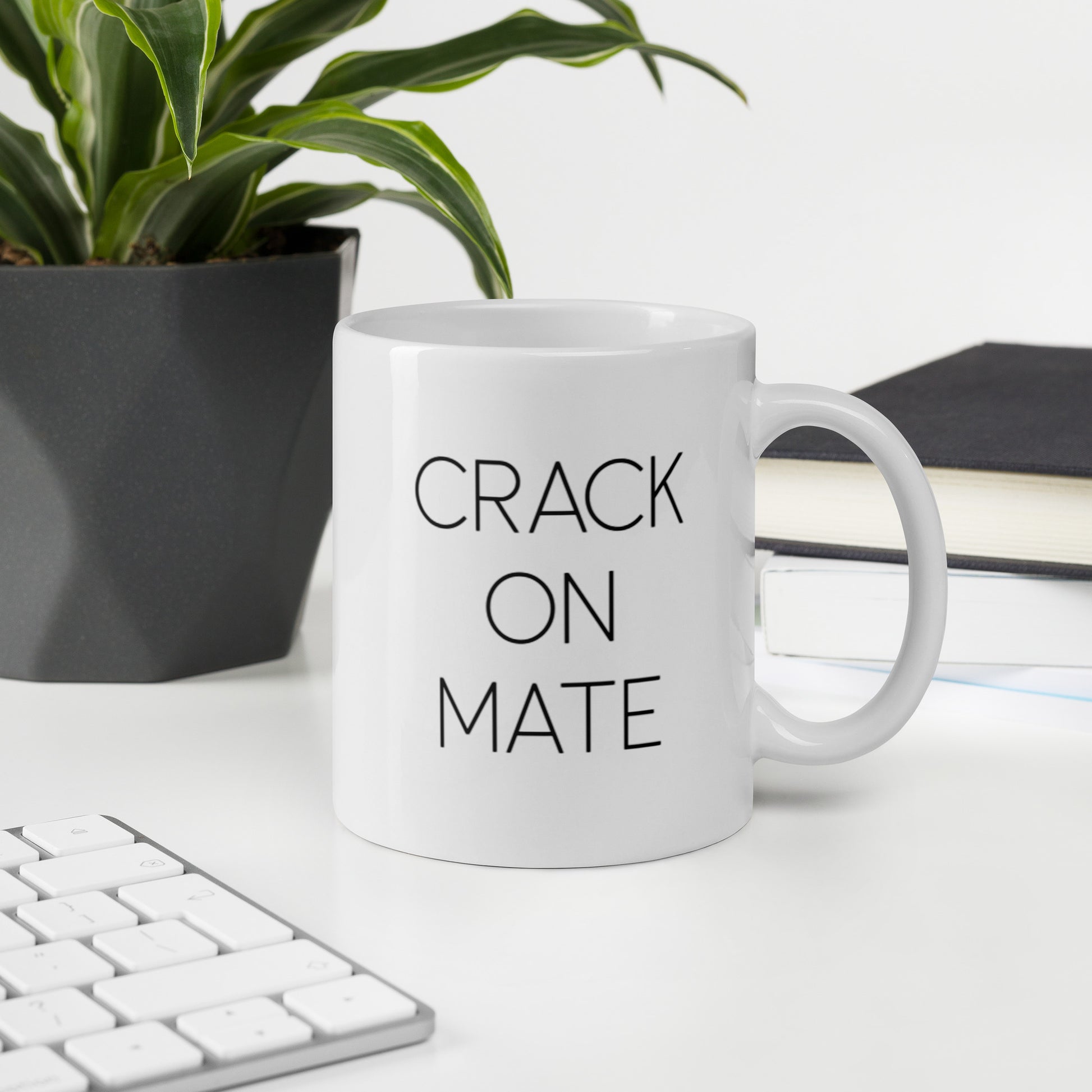 A glossy white ceramic mug with thin black all-caps text that reads 'CRACK ON MATE', sits upright on a white table. In the background, there's a potted plant with downward-curving leaves, resembling a snake plant, in a dark grey pot. Accompanying the plant is a stack of books. A corner of a keyboard is partially visible in the foreground, slightly out of focus, adding a subtle hint of workspace ambiance.