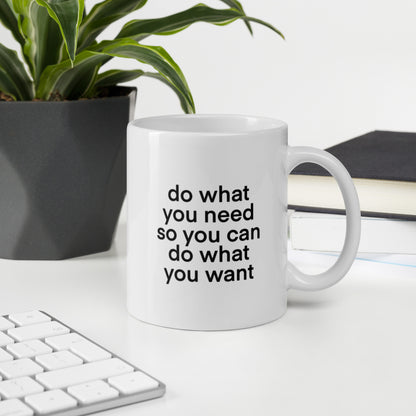 A glossy white ceramic mug with lowercase bolded black sans serif font that reads 'do what you need so you can do what you want', sits upright on a white table. In the background, there's a potted plant with downward-curving leaves, resembling a snake plant, in a dark grey pot. Accompanying the plant is a stack of books. A corner of a keyboard is partially visible in the foreground, slightly out of focus, adding a subtle hint of workspace ambiance.