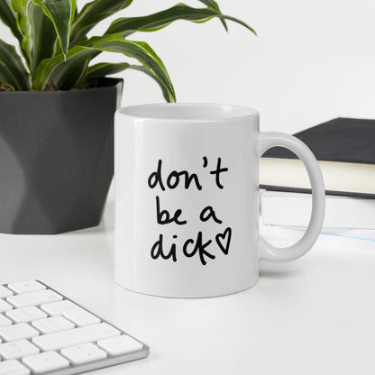 A glossy white ceramic mug with bold handwritten-style font that reads 'don't be a dick [ hand-drawn heart ]', sits upright on a white table. In the background, there's a potted plant with downward-curving leaves, resembling a snake plant, in a dark grey pot. Accompanying the plant is a stack of books. A corner of a keyboard is partially visible in the foreground, slightly out of focus, adding a subtle hint of workspace ambiance.