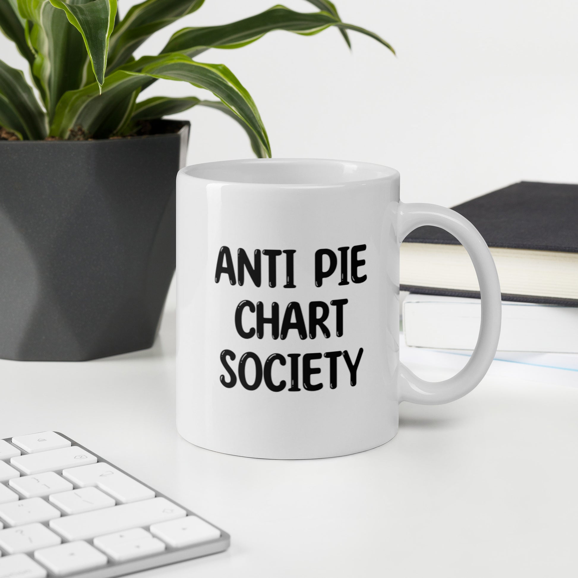 A glossy white ceramic mug with all-uppercase black bubble text that reads 'ANTI PIE CHART SOCIETY', sits upright on a white table. In the background, there's a potted plant with downward-curving leaves, resembling a snake plant, in a dark grey pot. Accompanying the plant is a stack of books. A corner of a keyboard is partially visible in the foreground, slightly out of focus, adding a subtle hint of workspace ambiance.