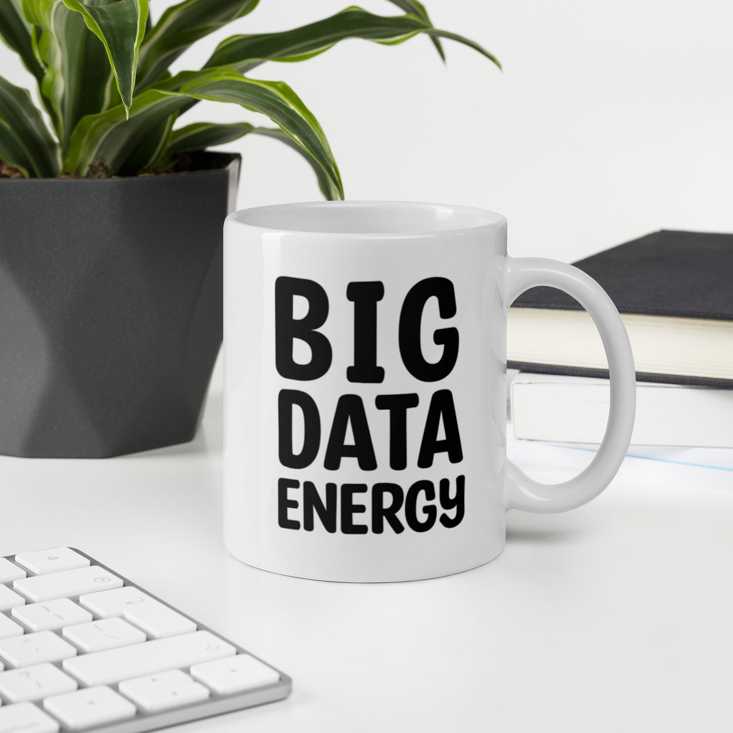 A glossy white ceramic mug with bolded black bubble text that reads 'BIG DATA ENERGY', sits upright on a white table. In the background, there's a potted plant with downward-curving leaves, resembling a snake plant, in a dark grey pot. Accompanying the plant is a stack of books. A corner of a keyboard is partially visible in the foreground, slightly out of focus, adding a subtle hint of workspace ambiance.