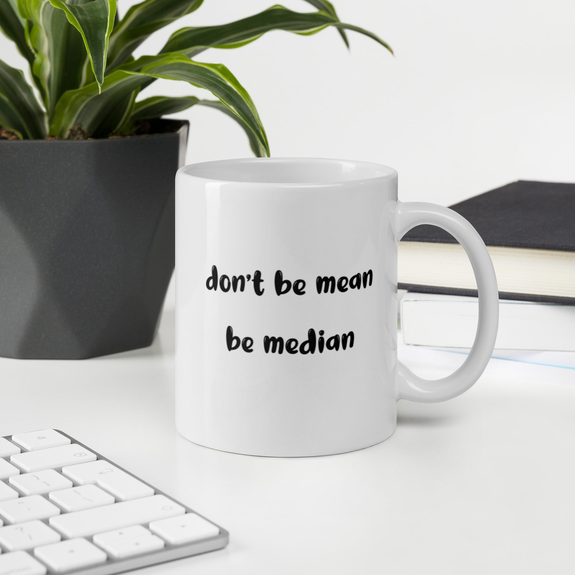 A glossy white ceramic mug with bold, lowercase black bubble font that reads 'don't be mean be median', sits upright on a white table. In the background, there's a potted plant with downward-curving leaves, resembling a snake plant, in a dark grey pot. Accompanying the plant is a stack of books. A corner of a keyboard is partially visible in the foreground, slightly out of focus, adding a subtle hint of workspace ambiance.