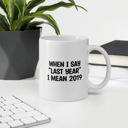 A glossy white ceramic mug with the text 'WHEN I SAY 'LAST YEAR' I MEAN 2019' in black bold uppercase font, sits upright on a white table. In the background, there's a potted plant with downward-curving leaves, resembling a snake plant, in a dark grey pot. Accompanying the plant is a stack of books. A corner of a keyboard is partially visible in the foreground, slightly out of focus, adding a subtle hint of workspace ambiance.