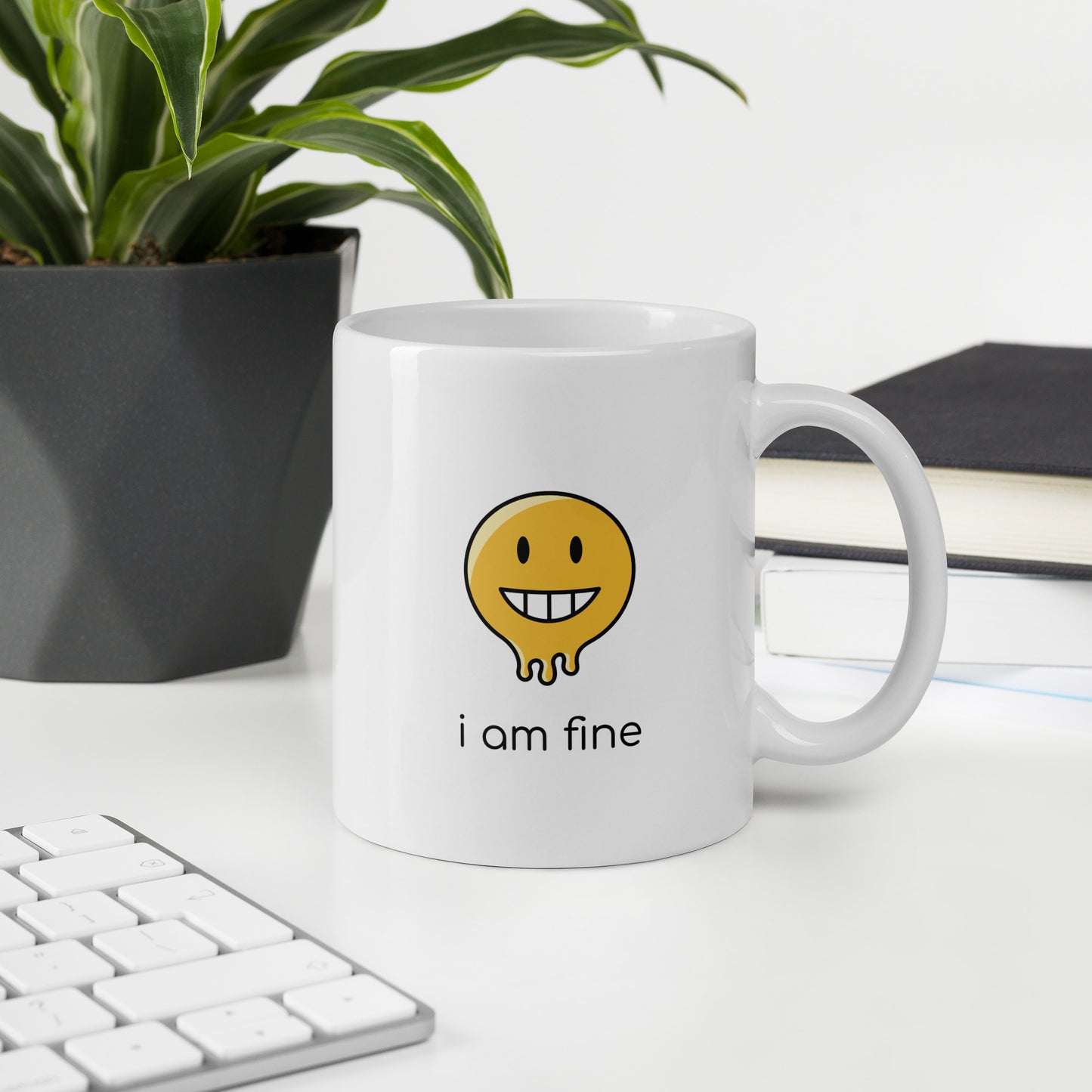 A glossy white ceramic mug with a graphic of a yellow smiley face (grinning) and melting at the bottom, accompanied by the caption 'i am fine' in small black sans serif text, sits upright on a white table. In the background, there's a potted plant with downward-curving leaves, resembling a snake plant, in a dark grey pot. Accompanying the plant is a stack of books. A corner of a keyboard is partially visible in the foreground, slightly out of focus, adding a subtle hint of workspace ambiance.