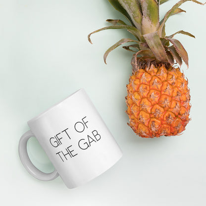 A glossy white ceramic mug with thin, all-caps black sans serif font that reads 'GIFT OF THE GAB', lies on its side next to a pineapple against a pastel light mint background.