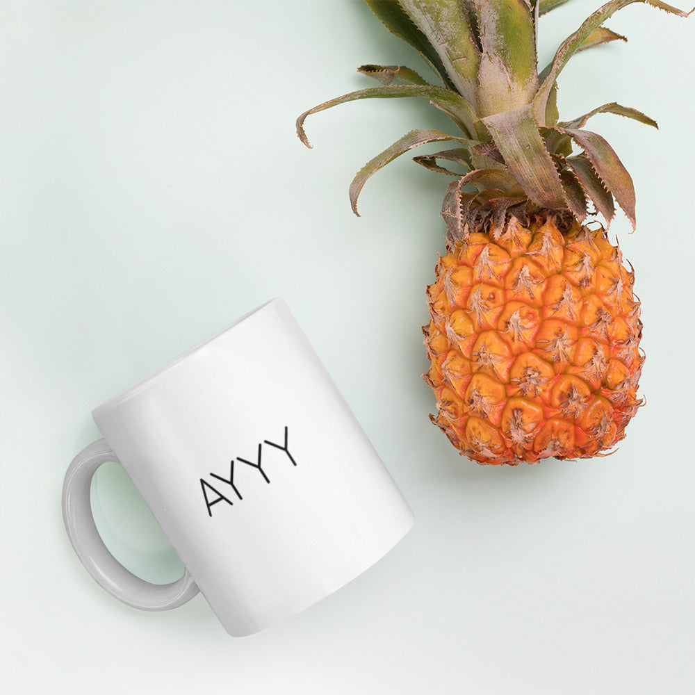 A glossy white ceramic mug with all-caps thin black text that reads 'AYYY', lies on its side next to a pineapple against a pastel light mint background.