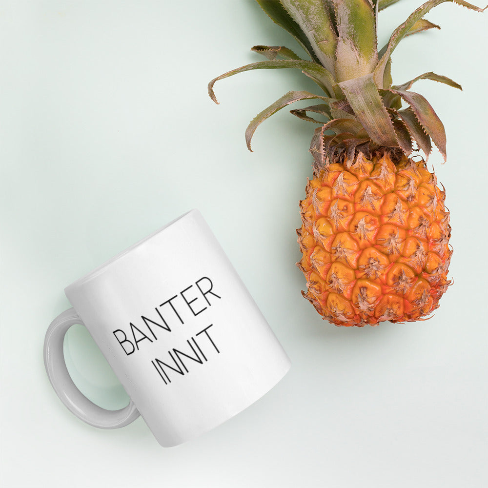 A glossy white ceramic mug with all-caps thin black text that reads 'BANTER INNIT', lies on its side next to a pineapple against a pastel light mint background.