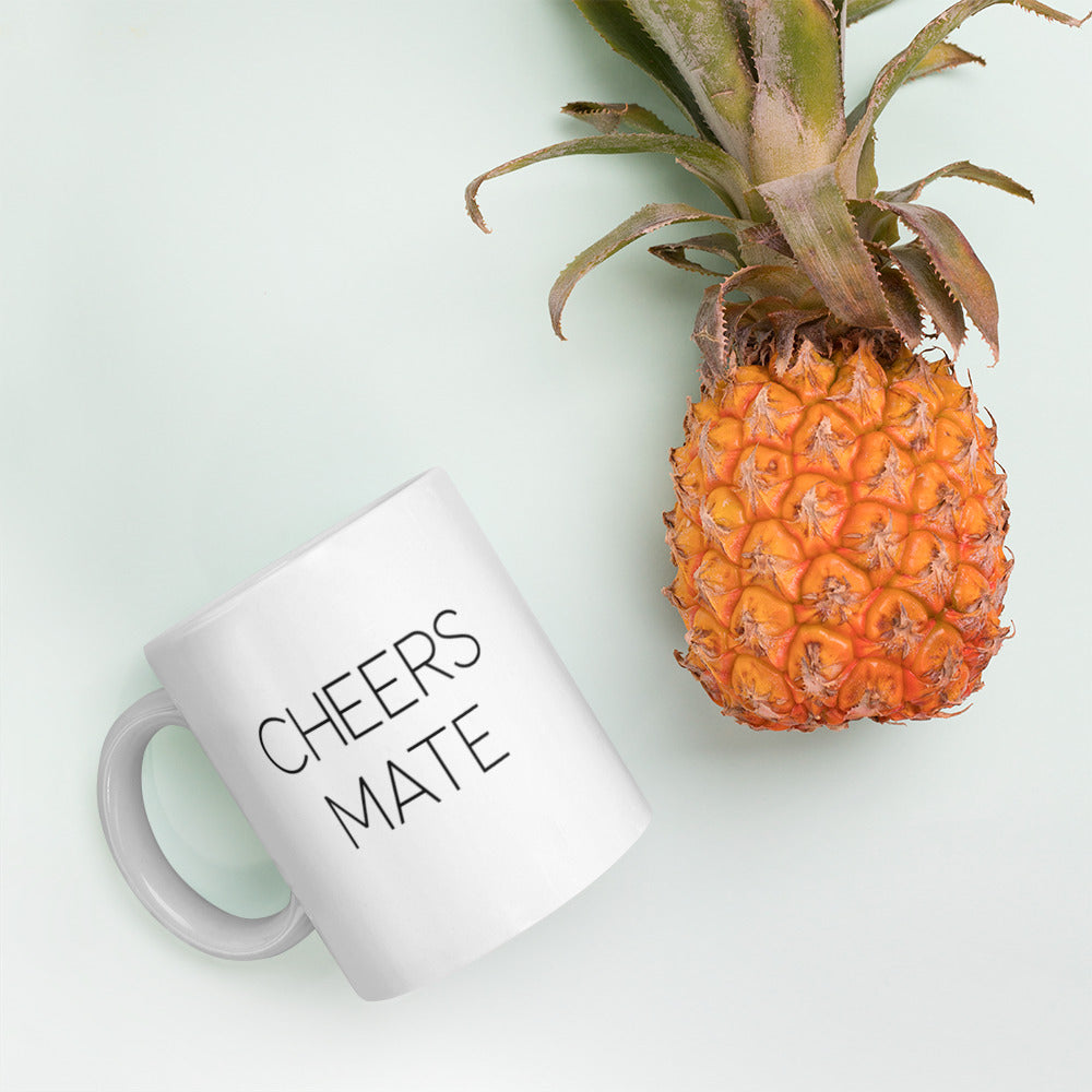 A glossy white ceramic mug with thin black all-caps text that reads 'CHEERS MATE', lies on its side next to a pineapple against a pastel light mint background.