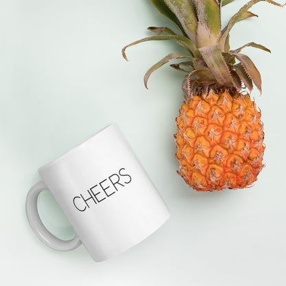 A glossy white ceramic mug with thin black all-caps text that reads 'CHEERS', lies on its side next to a pineapple against a pastel light mint background.