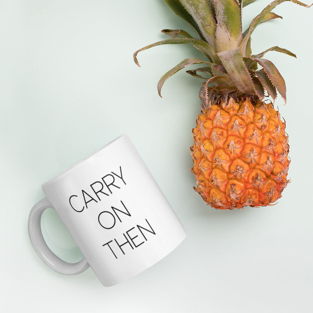 A glossy white ceramic mug with thin black all-caps text that reads 'CARRY ON THEN', lies on its side next to a pineapple against a pastel light mint background.