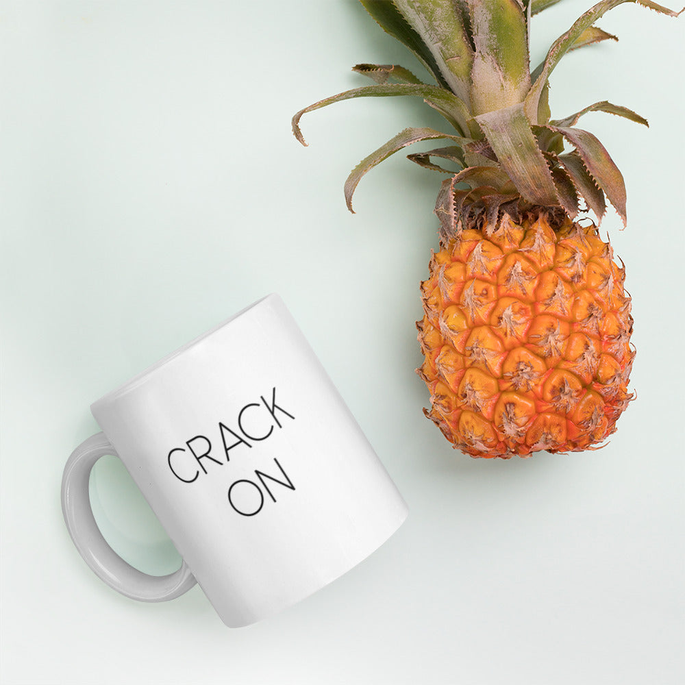 A glossy white ceramic mug with thin black all-caps text that reads 'CRACK ON', lies on its side next to a pineapple against a pastel light mint background.