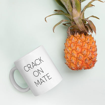 A glossy white ceramic mug with thin black all-caps text that reads 'CRACK ON MATE', lies on its side next to a pineapple against a pastel light mint background.