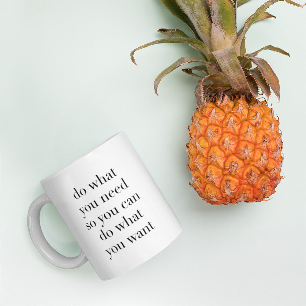 A glossy white ceramic mug with lowercase black serif font that reads 'do what you need so you can do what you want', lies on its side next to a pineapple against a pastel light mint background.