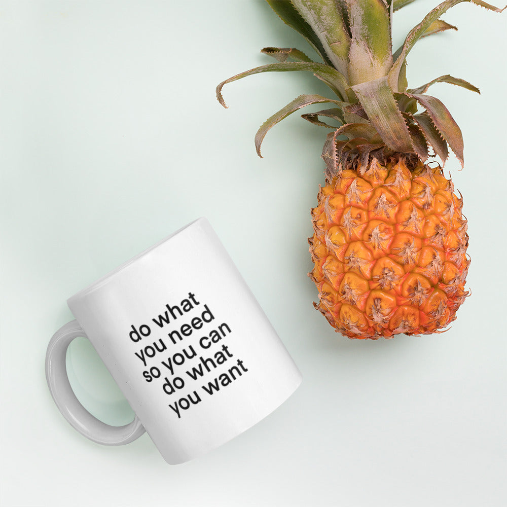 A glossy white ceramic mug with lowercase bolded black sans serif font that reads 'do what you need so you can do what you want', lies on its side next to a pineapple against a pastel light mint background.