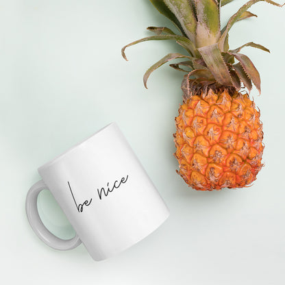 A glossy white ceramic mug with script text that reads 'be nice', lies on its side next to a pineapple against a pastel light mint background.