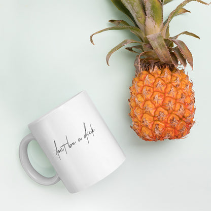 A glossy white ceramic mug with lowercase script font that reads 'don't be a dick', lies on its side next to a pineapple against a pastel light mint background.