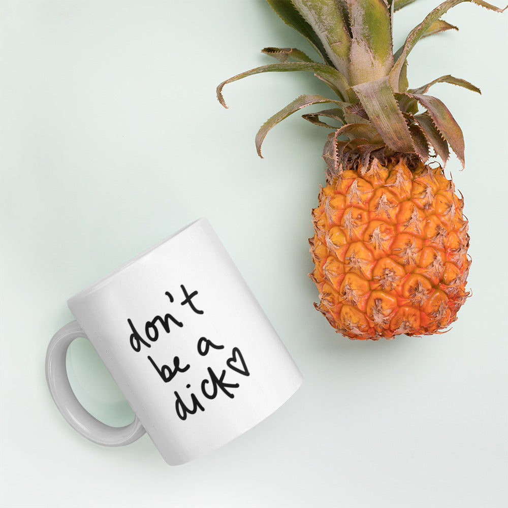 A glossy white ceramic mug with bold handwritten-style font that reads 'don't be a dick [ hand-drawn heart ]', lies on its side next to a pineapple against a pastel light mint background.