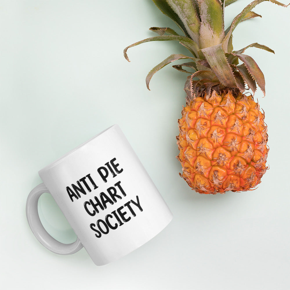 A glossy white ceramic mug with all-uppercase black bubble text that reads 'ANTI PIE CHART SOCIETY', lies on its side next to a pineapple against a pastel light mint background.