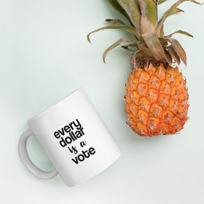 A glossy white ceramic mug with bold, lowercase black sans serif font that reads 'every dollar is a vote', with 'is a' in black handwritten script font, lies on its side next to a pineapple against a pastel light mint background.