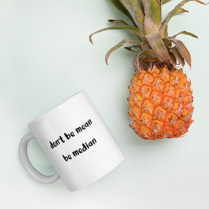 A glossy white ceramic mug with bold, lowercase black bubble font that reads 'don't be mean be median', lies on its side next to a pineapple against a pastel light mint background.