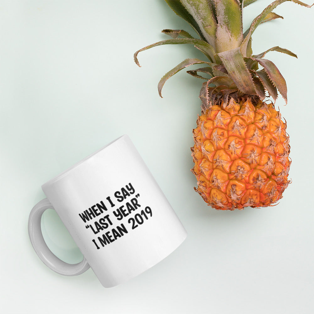 A glossy white ceramic mug with the text 'WHEN I SAY 'LAST YEAR' I MEAN 2019' in black bold uppercase font, lies on its side next to a pineapple against a pastel light mint background.