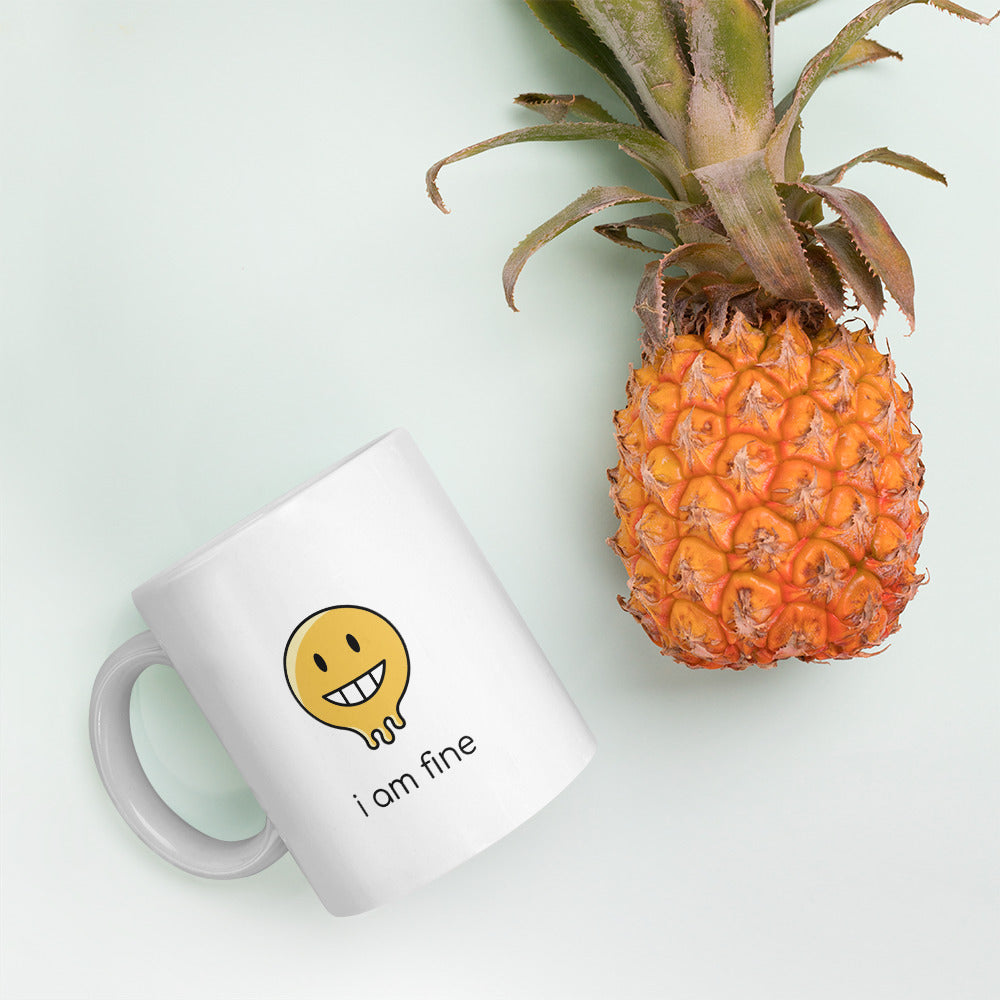 A glossy white ceramic mug with a graphic of a yellow smiley face (grinning) and melting at the bottom, accompanied by the caption 'i am fine' in small black sans serif text, lies on its side next to a pineapple against a pastel light mint background.