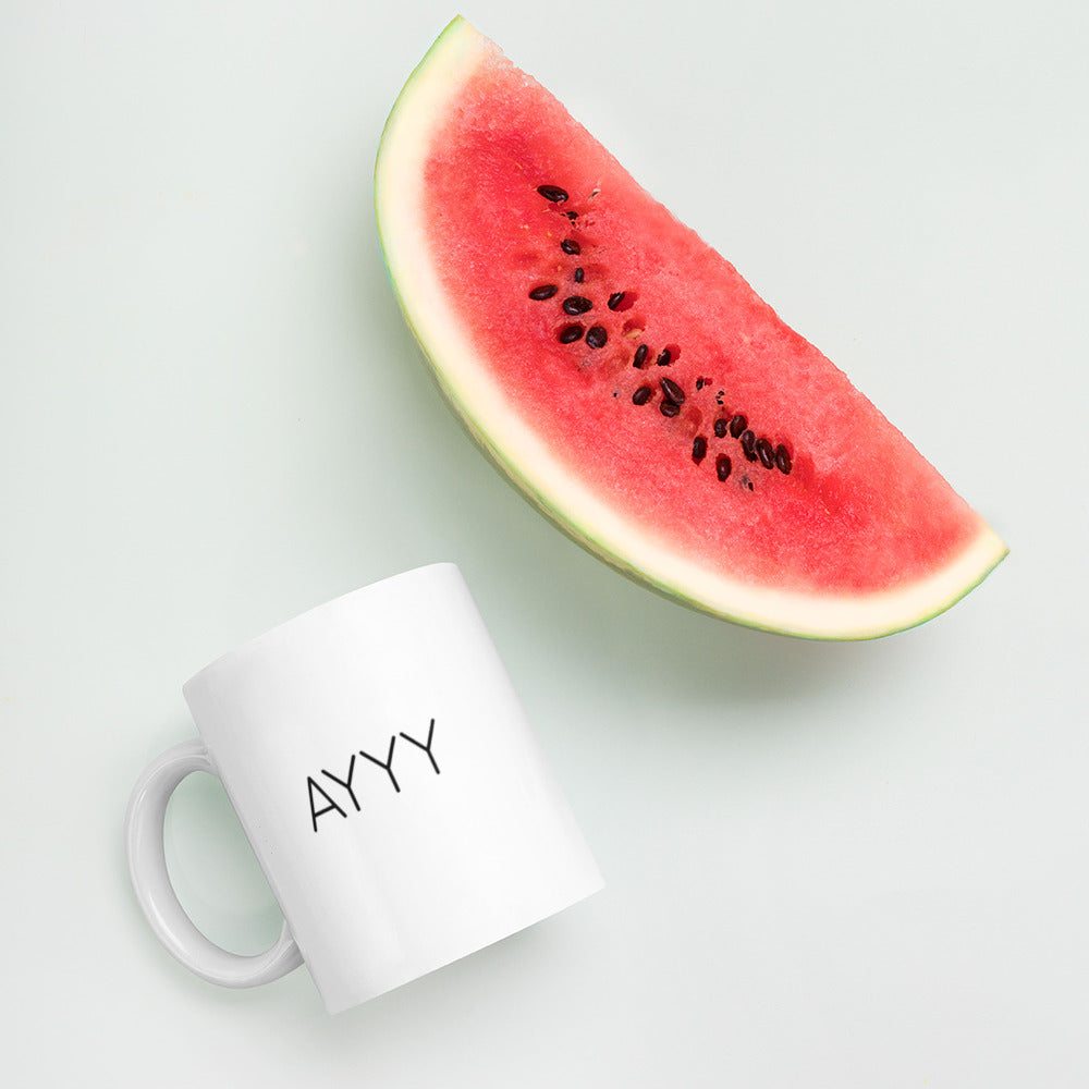 A glossy white ceramic mug with all-caps thin black text that reads 'AYYY', lies on its side next to a slice of watermelon against a pastel light mint background.