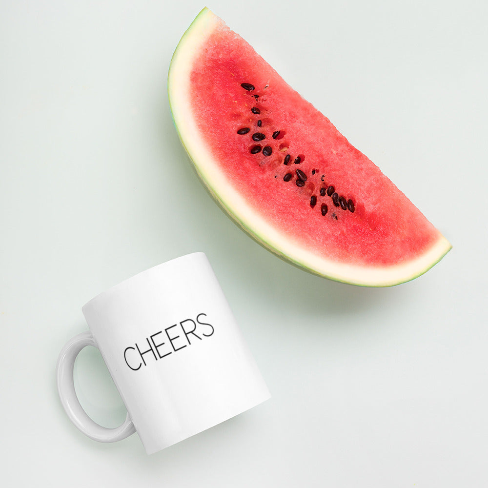 A glossy white ceramic mug with thin black all-caps text that reads 'CHEERS', lies on its side next to a slice of watermelon against a pastel light mint background.