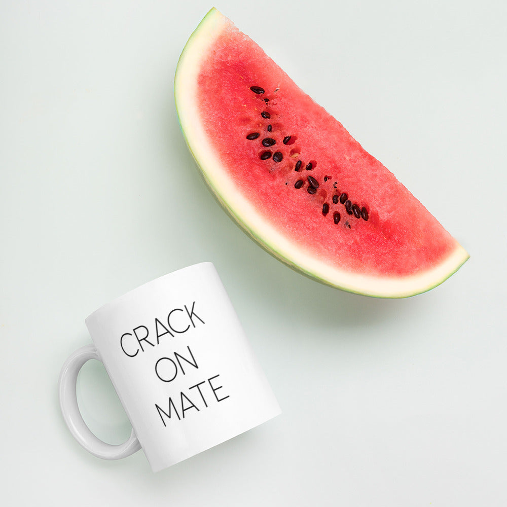 A glossy white ceramic mug with thin black all-caps text that reads 'CRACK ON MATE', lies on its side next to a slice of watermelon against a pastel light mint background.