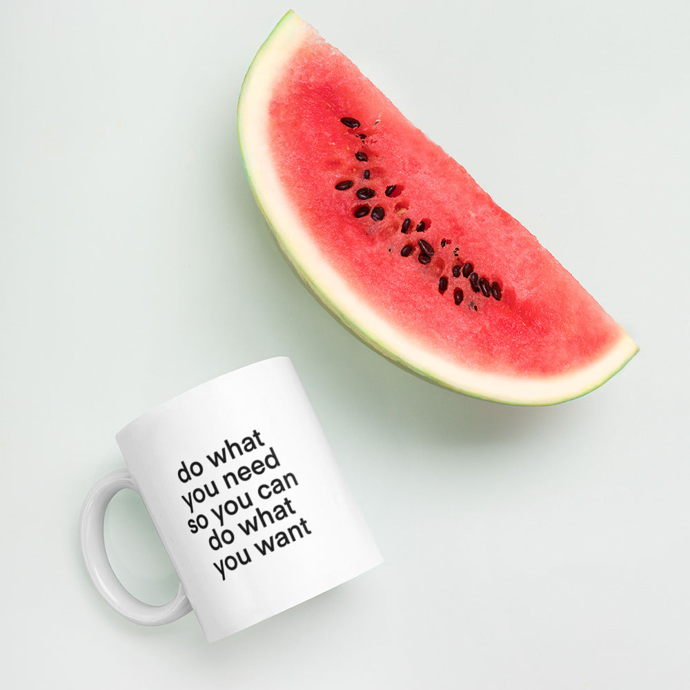 A glossy white ceramic mug with lowercase bolded black sans serif font that reads 'do what you need so you can do what you want', lies on its side next to a slice of watermelon against a pastel light mint background.