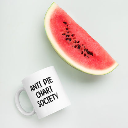 A glossy white ceramic mug with all-uppercase black bubble text that reads 'ANTI PIE CHART SOCIETY', lies on its side next to a slice of watermelon against a pastel light mint background.