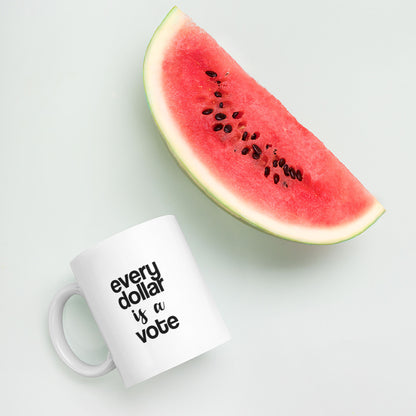 A glossy white ceramic mug with bold, lowercase black sans serif font that reads 'every dollar is a vote', with 'is a' in black handwritten script font, lies on its side next to a slice of watermelon against a pastel light mint background.