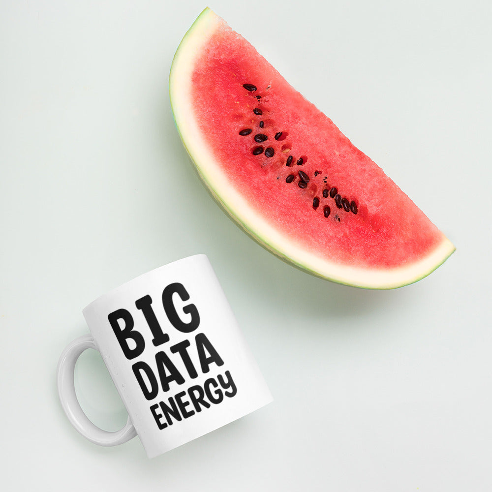 A glossy white ceramic mug with bolded black bubble text that reads 'BIG DATA ENERGY', lies on its side next to a slice of watermelon against a pastel light mint background.