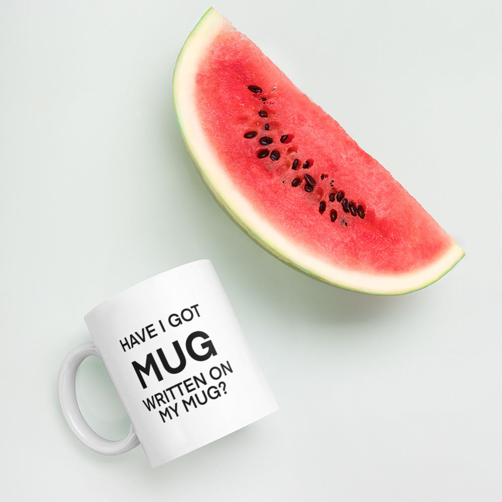 A glossy white ceramic mug with black bolded sans serif text that reads 'HAVE I GOT MUG WRITTEN ON MY MUG?', where the first 'MUG' is in bigger font size, lies on its side next to a slice of watermelon against a pastel light mint background.