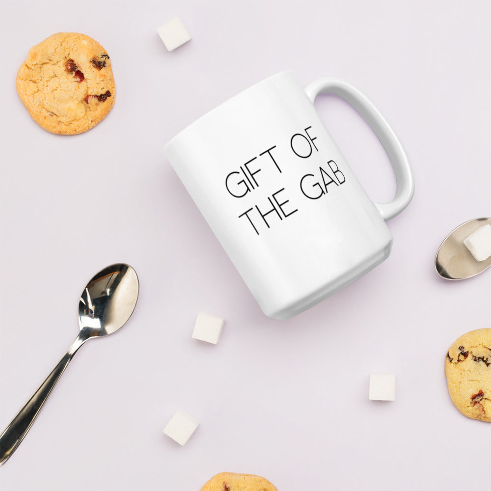 A glossy white ceramic mug with thin, all-caps black sans serif font that reads 'GIFT OF THE GAB', lies on its side against a very pale lavender blush background. Surrounding the mug are a few chocolate chip cookies, five cubes of sugar, and two spoons.