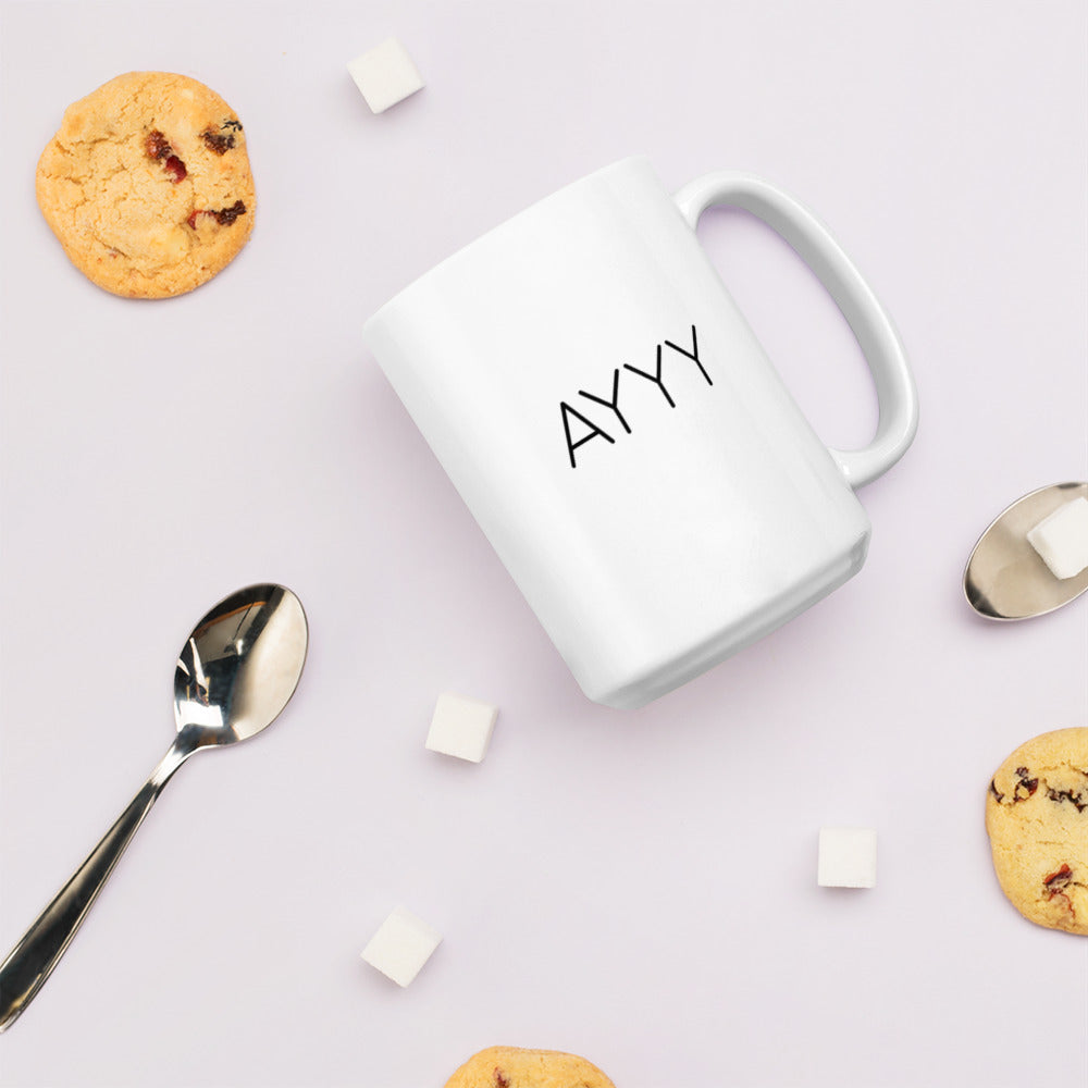 A glossy white ceramic mug with all-caps thin black text that reads 'AYYY', lies on its side against a very pale lavender blush background. Surrounding the mug are a few chocolate chip cookies, five cubes of sugar, and two spoons.