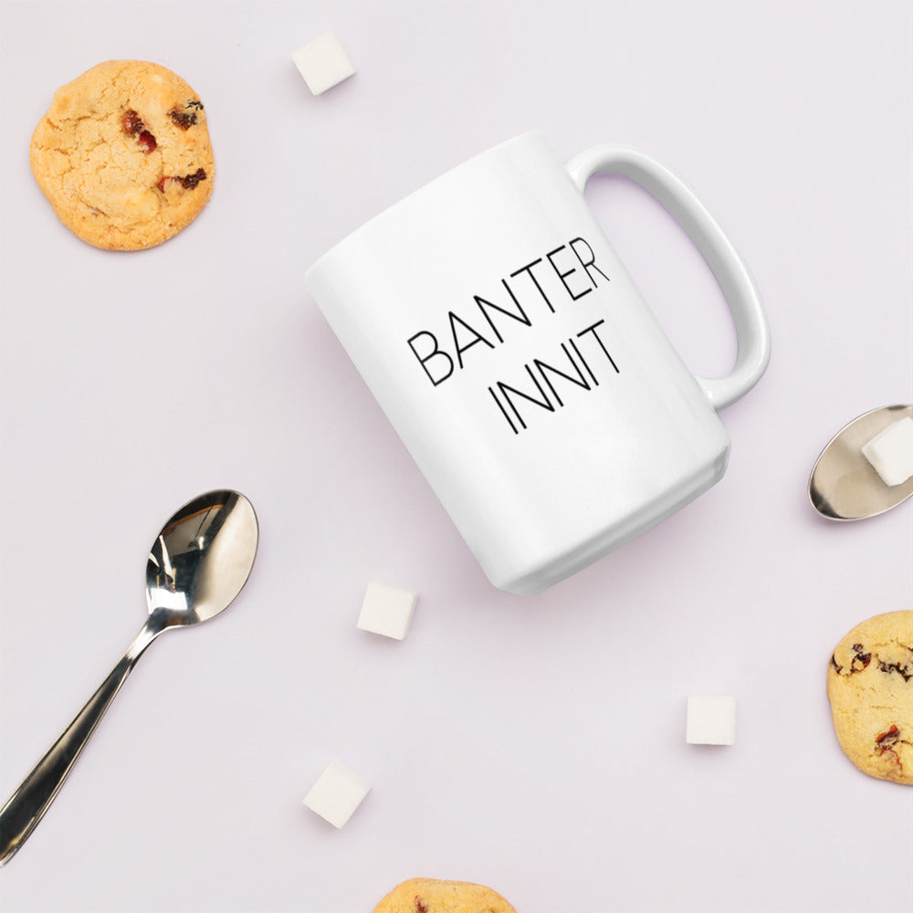 A glossy white ceramic mug with all-caps thin black text that reads 'BANTER INNIT', lies on its side against a very pale lavender blush background. Surrounding the mug are a few chocolate chip cookies, five cubes of sugar, and two spoons.