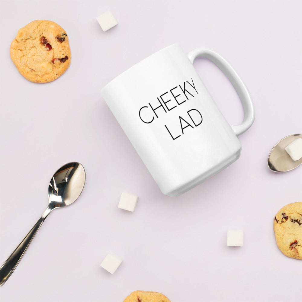 A glossy white ceramic mug with thin black all-caps text that reads 'CHEEKY LAD', lies on its side against a very pale lavender blush background. Surrounding the mug are a few chocolate chip cookies, five cubes of sugar, and two spoons.