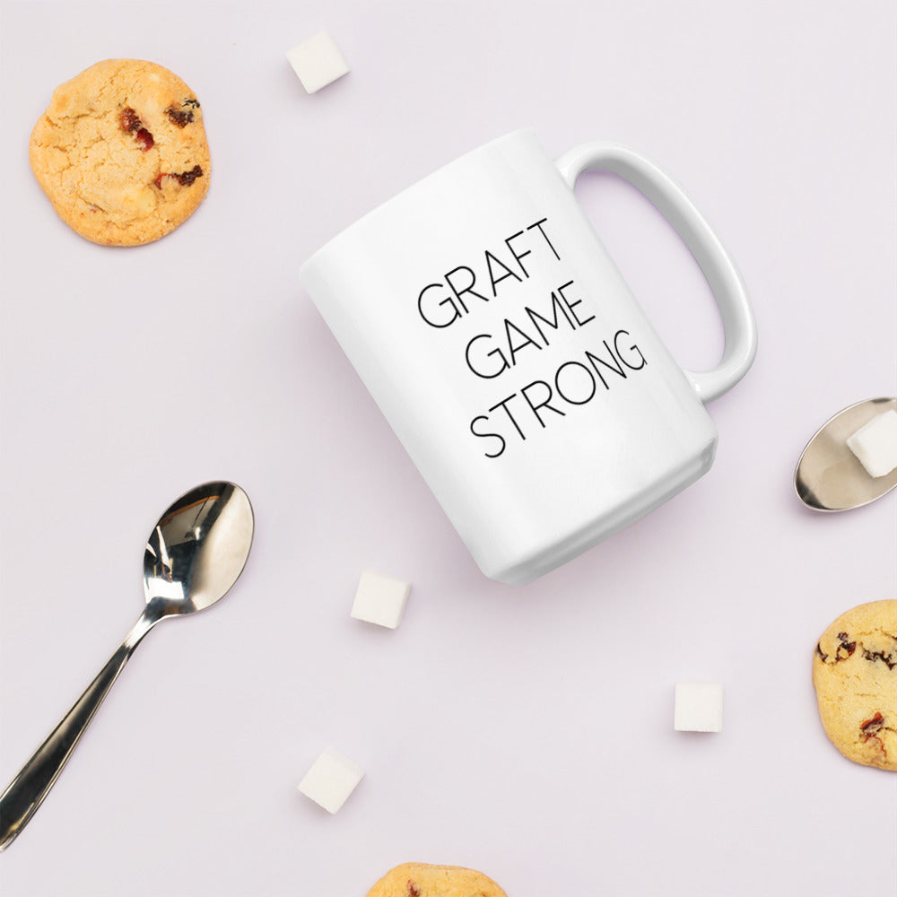 A glossy white ceramic mug with thin, all-caps black sans serif font that reads 'GRAFT GAME STRONG', lies on its side against a very pale lavender blush background. Surrounding the mug are a few chocolate chip cookies, five cubes of sugar, and two spoons.