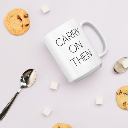 A glossy white ceramic mug with thin black all-caps text that reads 'CARRY ON THEN', lies on its side against a very pale lavender blush background. Surrounding the mug are a few chocolate chip cookies, five cubes of sugar, and two spoons.